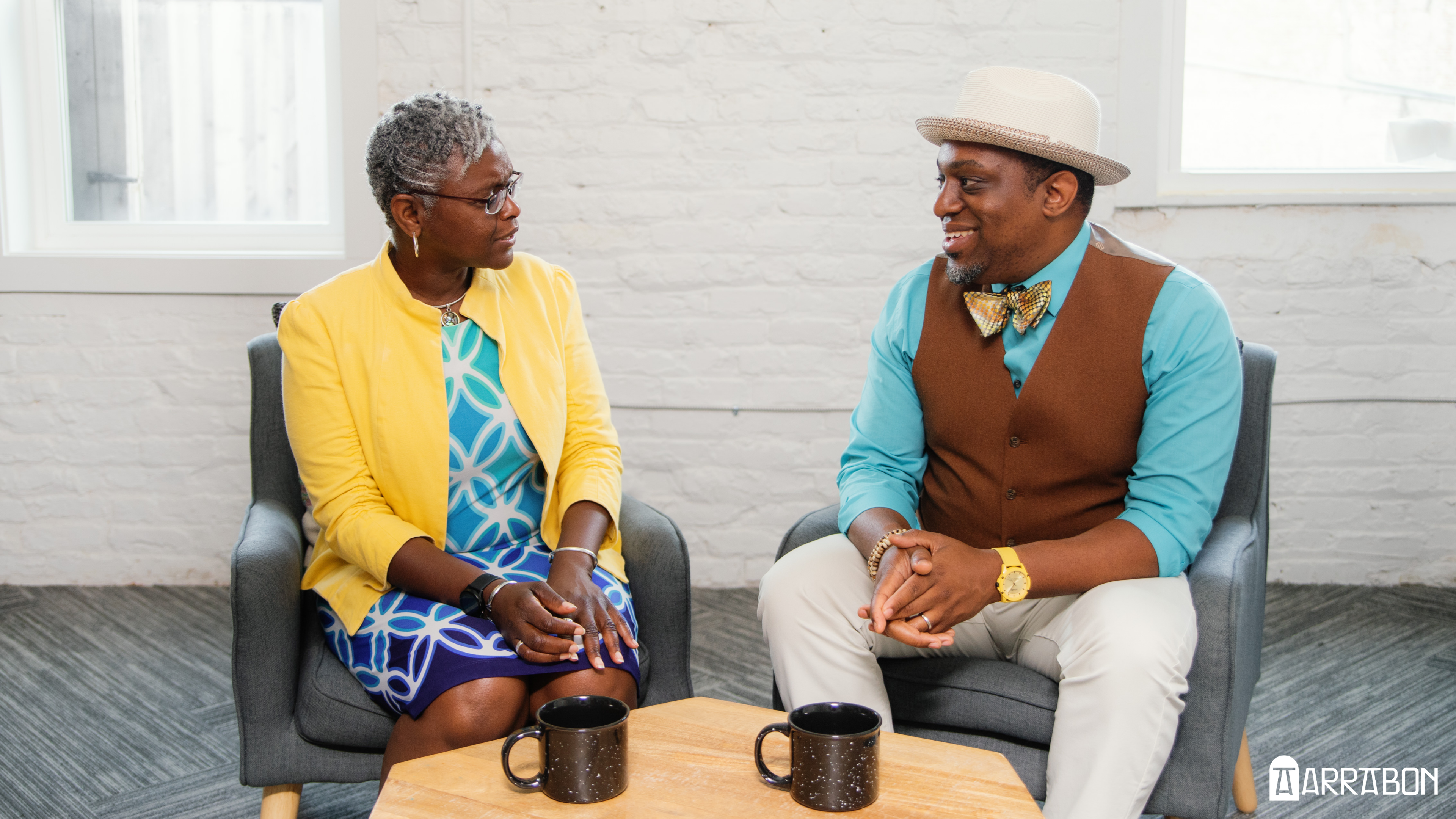 Abigail, wearing a yellow jacket and blue-green sheath dress, faces David who is wearing a tan hat, teal shirt, and brown vest. There’s a coffee table in front of them with two mugs.