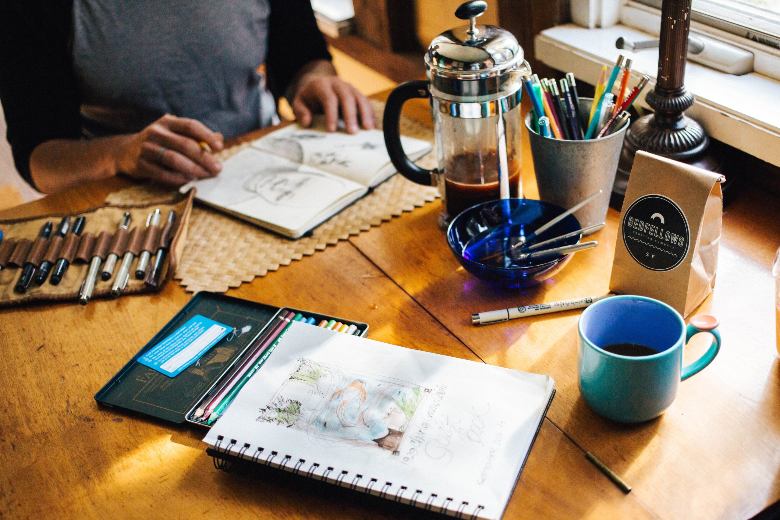 A person sits at a wood table with a journal open and various colored pencils and art supplies. A french press and coffee also sits on the table